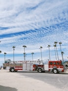 A fire truck from the Los Angeles Fire Department is parked on a sandy beach area. The background features tall palm trees and a partly cloudy blue sky, suggesting a coastal location.