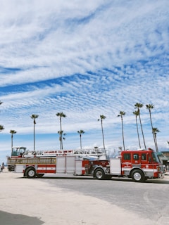 A fire truck from the Los Angeles Fire Department is parked on a sandy beach area. The background features tall palm trees and a partly cloudy blue sky, suggesting a coastal location.