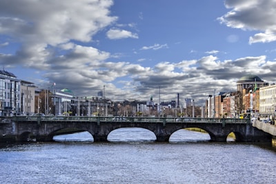 A river flowing beneath a bridge with multiple arches is flanked by urban architecture. The sky above features scattered clouds with patches of blue, while the cityscape extends into the distance with various buildings, some topped with green domes.