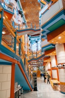 A modern and vibrant interior with colorful architectural features including blue, orange, and green elements. The design includes angular lines, geometric patterns, and a mix of materials. Several people are walking through the space, which has multiple levels connected by walkways and stairs. The ceiling is adorned with a unique pattern, adding to the dynamic visual effect.