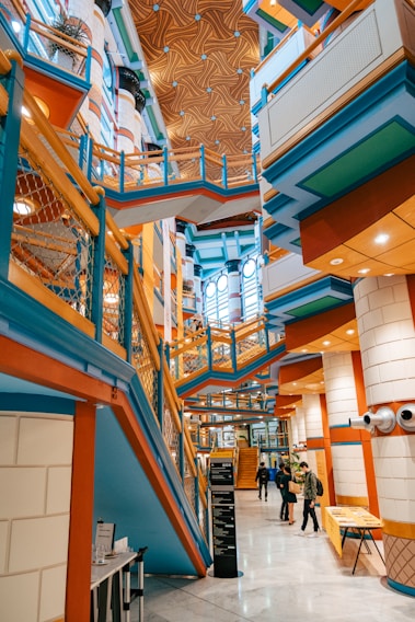 A modern and vibrant interior with colorful architectural features including blue, orange, and green elements. The design includes angular lines, geometric patterns, and a mix of materials. Several people are walking through the space, which has multiple levels connected by walkways and stairs. The ceiling is adorned with a unique pattern, adding to the dynamic visual effect.