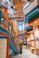 A modern and vibrant interior with colorful architectural features including blue, orange, and green elements. The design includes angular lines, geometric patterns, and a mix of materials. Several people are walking through the space, which has multiple levels connected by walkways and stairs. The ceiling is adorned with a unique pattern, adding to the dynamic visual effect.