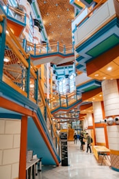 A modern and vibrant interior with colorful architectural features including blue, orange, and green elements. The design includes angular lines, geometric patterns, and a mix of materials. Several people are walking through the space, which has multiple levels connected by walkways and stairs. The ceiling is adorned with a unique pattern, adding to the dynamic visual effect.