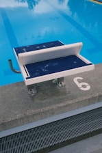 A swimming pool with clear blue water is visible, featuring a diving platform numbered '6' and a handrail on its side. The platform is positioned at the edge of the pool, which has lane markings visible underwater.