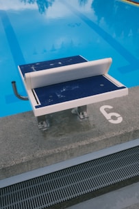 A swimming pool with clear blue water is visible, featuring a diving platform numbered '6' and a handrail on its side. The platform is positioned at the edge of the pool, which has lane markings visible underwater.