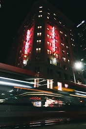 A nighttime city scene featuring a tall building illuminated with bright red lights on the facade spelling 'GOWINGS'. The windows of the building show varied lighting, suggesting occupied floors. A motion-blurred vehicle, possibly a tram or bus, passes by in the foreground, with the word 'MECCA' visible on a storefront.