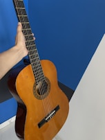 A hand is holding a classical acoustic guitar against a blue and white background. The guitar has a wooden body with six strings and a detailed rosette around the soundhole.
