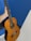 A hand is holding a classical acoustic guitar against a blue and white background. The guitar has a wooden body with six strings and a detailed rosette around the soundhole.