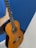 A hand is holding a classical acoustic guitar against a blue and white background. The guitar has a wooden body with six strings and a detailed rosette around the soundhole.