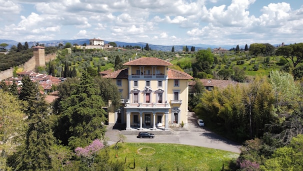 A large, elegant villa with a yellow facade and red-tiled roof is set among lush greenery. The villa is surrounded by tall trees and shrubs, with a driveway leading up to it. In the background, rolling hills with scattered buildings and a stone wall add to the picturesque landscape. The sky above is filled with fluffy white clouds.