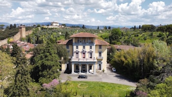 A large, elegant villa with a yellow facade and red-tiled roof is set among lush greenery. The villa is surrounded by tall trees and shrubs, with a driveway leading up to it. In the background, rolling hills with scattered buildings and a stone wall add to the picturesque landscape. The sky above is filled with fluffy white clouds.