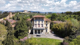 A large, elegant villa with a yellow facade and red-tiled roof is set among lush greenery. The villa is surrounded by tall trees and shrubs, with a driveway leading up to it. In the background, rolling hills with scattered buildings and a stone wall add to the picturesque landscape. The sky above is filled with fluffy white clouds.