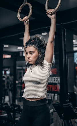 A person with curly hair is exercising in a gym, holding onto gymnastic rings. They are wearing a striped top and dark leggings. The background features gym equipment and motivational posters.