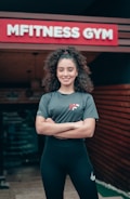 A woman with curly hair stands confidently in front of a gym entrance, with a sign reading 'MFITNESS GYM' above. She is wearing a sporty outfit consisting of a grey T-shirt and black leggings, and has her arms crossed while smiling.