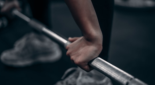 A close-up of hands gripping a barbell, with a focus on the knurled metal surface of the bar. The background is slightly blurred, showing a pair of sneakers on a dark floor, indicating a gym or fitness setting.