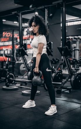 A woman with curly hair is performing a workout with a kettlebell in a gym environment. She is wearing a striped shirt, black leggings, and white sneakers. The gym is equipped with weights, benches, and other fitness equipment.