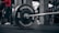 A close-up view of a barbell with weight plates in a gym setting. The weight plates read 'Crossmaxx' and have a weight of 5 kg. There is an individual in red athletic wear in the background and gym equipment is visible.