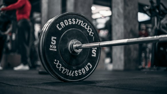 A close-up view of a barbell with weight plates in a gym setting. The weight plates read 'Crossmaxx' and have a weight of 5 kg. There is an individual in red athletic wear in the background and gym equipment is visible.
