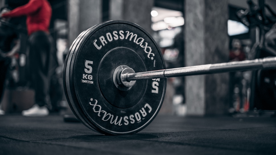 A close-up view of a barbell with weight plates in a gym setting. The weight plates read 'Crossmaxx' and have a weight of 5 kg. There is an individual in red athletic wear in the background and gym equipment is visible.