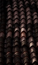 Rows of dark, weathered roof tiles with a textured surface, some displaying signs of moss or dirt accumulation, casting shadows and creating a rhythmic pattern.