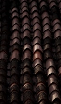 Rows of dark, weathered roof tiles with a textured surface, some displaying signs of moss or dirt accumulation, casting shadows and creating a rhythmic pattern.