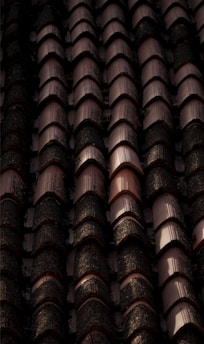 Rows of dark, weathered roof tiles with a textured surface, some displaying signs of moss or dirt accumulation, casting shadows and creating a rhythmic pattern.