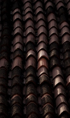 Rows of dark, weathered roof tiles with a textured surface, some displaying signs of moss or dirt accumulation, casting shadows and creating a rhythmic pattern.