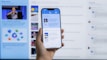 A hand holding a smartphone displaying a news website, with a blurred large computer screen in the background also showing similar content. The webpage on the phone features articles with images and text.