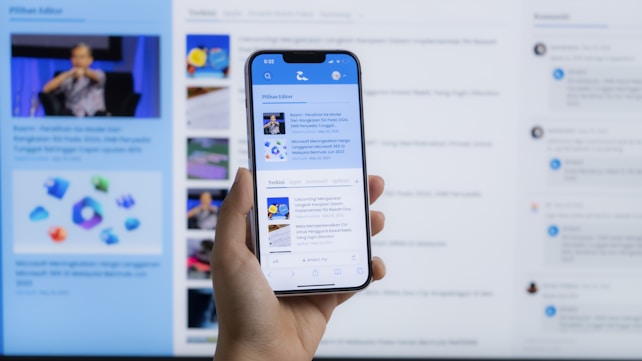 A hand holding a smartphone displaying a news website, with a blurred large computer screen in the background also showing similar content. The webpage on the phone features articles with images and text.