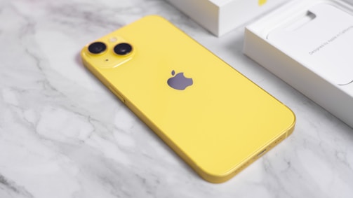 A yellow smartphone lies on a marble surface with its back facing up, displaying the Apple logo. The phone has dual rear cameras on the top left corner. In the background, there is a white box, likely the phone's packaging, partially visible.