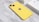 A yellow smartphone lies on a marble surface with its back facing up, displaying the Apple logo. The phone has dual rear cameras on the top left corner. In the background, there is a white box, likely the phone's packaging, partially visible.