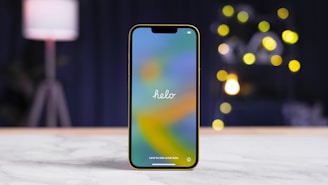 A smartphone with a colorful screen displaying the word 'helo' is positioned upright on a white marble surface. The background features a blurred scene with a contemporary lamp and several round, bokeh-style lights, giving a cozy ambiance.