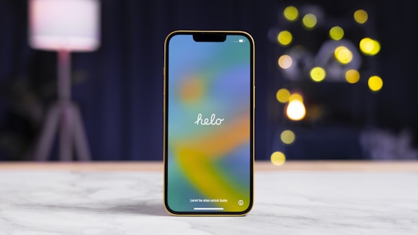 A smartphone with a colorful screen displaying the word 'helo' is positioned upright on a white marble surface. The background features a blurred scene with a contemporary lamp and several round, bokeh-style lights, giving a cozy ambiance.