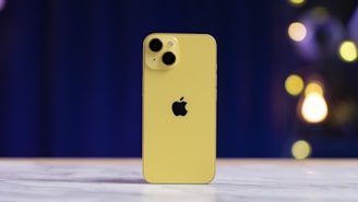 A yellow smartphone standing upright on a marble surface, featuring a dual-camera setup and an apple logo. The background is blurred with blue and yellow lights creating a bokeh effect.