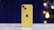 A yellow smartphone standing upright on a marble surface, featuring a dual-camera setup and an apple logo. The background is blurred with blue and yellow lights creating a bokeh effect.