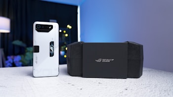 A sleek white smartphone with a prominent black camera module and futuristic design elements is placed on a textured surface. Beside it, a black box with the logo 'Republic of Gamers' is positioned. The background includes a softly lit lamp, plants, and a faintly glowing light, creating a modern and tech-focused atmosphere.