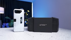 A sleek white smartphone with a prominent black camera module and futuristic design elements is placed on a textured surface. Beside it, a black box with the logo 'Republic of Gamers' is positioned. The background includes a softly lit lamp, plants, and a faintly glowing light, creating a modern and tech-focused atmosphere.