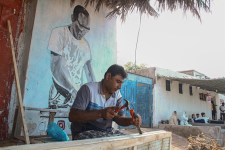 A man is focused on a woodworking task, using a hammer and chisel. Behind him, a mural of another person is painted on the wall. Nearby, several people are seated in a shaded area. The setting appears to be outdoors with simple structures and palm leaves forming part of the roof.
