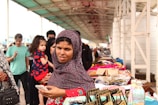 A bustling market scene with a woman in the foreground wearing a patterned headscarf and holding a smartphone. She is standing near a stall displaying various textiles and colorful bags. In the background, more people are visible, including a person holding a child. The market is set under a covered walkway with a busy atmosphere.