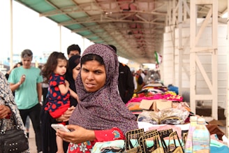 A bustling market scene with a woman in the foreground wearing a patterned headscarf and holding a smartphone. She is standing near a stall displaying various textiles and colorful bags. In the background, more people are visible, including a person holding a child. The market is set under a covered walkway with a busy atmosphere.