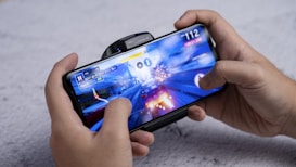 A person is holding a smartphone horizontally, playing a racing video game. The screen shows a high-speed race scene with a car emitting sparks. The hands are gripping the device tightly, indicating focus and engagement in the game.