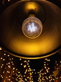 A close-up view of a dangling incandescent light bulb with a textured yellow lamp shade above it. Below, strings of small, warm white fairy lights create a bokeh effect against a dark background. The setting seems to evoke a festive or cozy atmosphere.