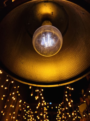 A close-up view of a dangling incandescent light bulb with a textured yellow lamp shade above it. Below, strings of small, warm white fairy lights create a bokeh effect against a dark background. The setting seems to evoke a festive or cozy atmosphere.