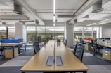 A modern, open-plan office space with long wooden desks and ergonomic chairs. The room is well-lit with natural light streaming through large windows. The ceiling features exposed ductwork and fluorescent lighting. The decor is minimal, with blue and orange accents on dividers and storage units.