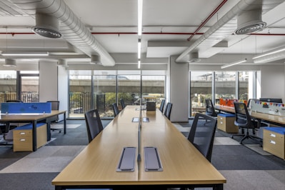 A modern, open-plan office space with long wooden desks and ergonomic chairs. The room is well-lit with natural light streaming through large windows. The ceiling features exposed ductwork and fluorescent lighting. The decor is minimal, with blue and orange accents on dividers and storage units.