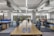 A modern, open-plan office space with long wooden desks and ergonomic chairs. The room is well-lit with natural light streaming through large windows. The ceiling features exposed ductwork and fluorescent lighting. The decor is minimal, with blue and orange accents on dividers and storage units.
