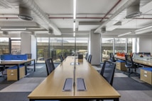 A modern, open-plan office space with long wooden desks and ergonomic chairs. The room is well-lit with natural light streaming through large windows. The ceiling features exposed ductwork and fluorescent lighting. The decor is minimal, with blue and orange accents on dividers and storage units.