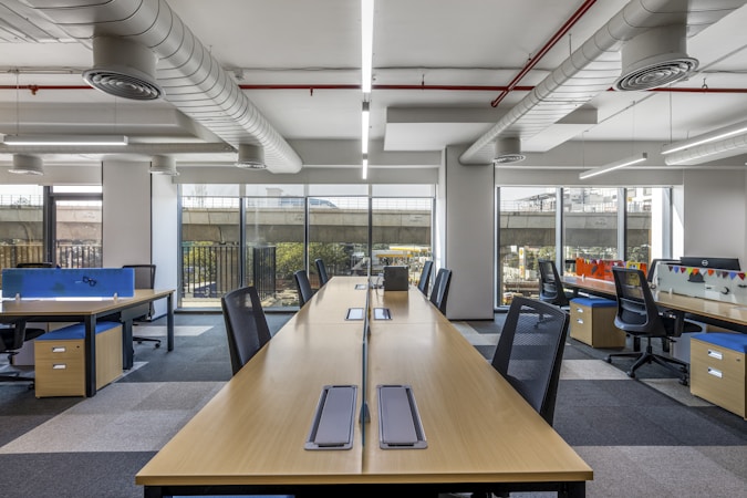 A modern, open-plan office space with long wooden desks and ergonomic chairs. The room is well-lit with natural light streaming through large windows. The ceiling features exposed ductwork and fluorescent lighting. The decor is minimal, with blue and orange accents on dividers and storage units.