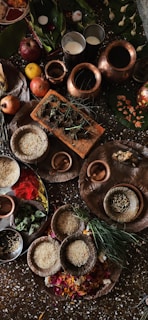 A collection of items, including copper vessels filled with liquids, plates containing rice, spices, and other ingredients, surrounded by fruits such as apples and bananas, and green leaves. The setting appears ceremonial or ritualistic, with a variety of natural elements and earthenware.