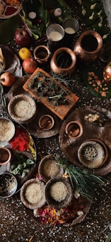 A collection of items, including copper vessels filled with liquids, plates containing rice, spices, and other ingredients, surrounded by fruits such as apples and bananas, and green leaves. The setting appears ceremonial or ritualistic, with a variety of natural elements and earthenware.